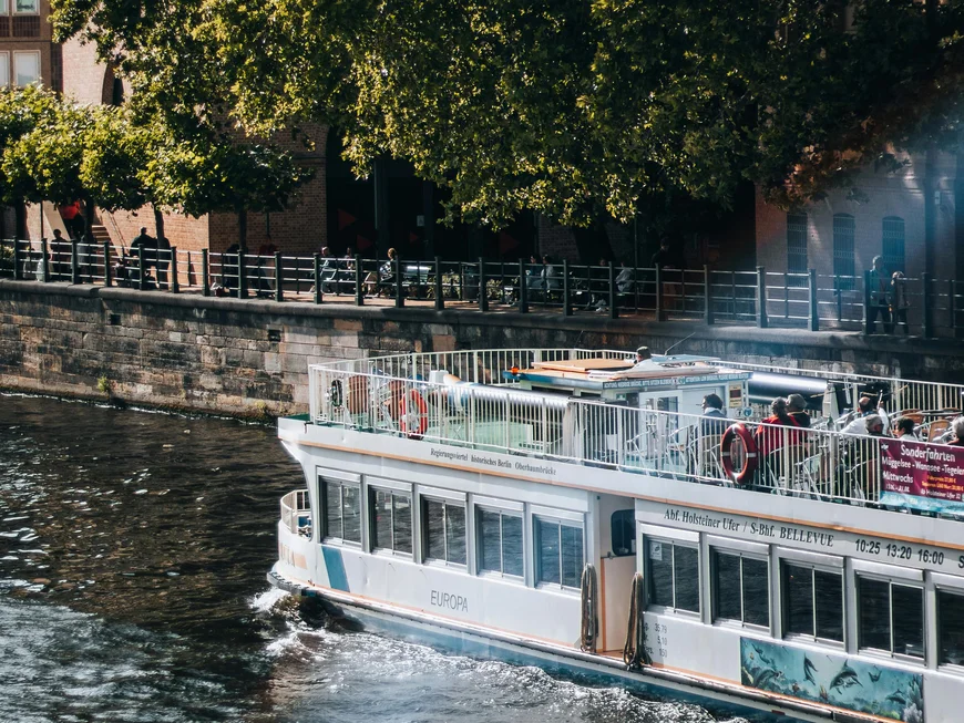 Berliner Sehenswürdigkeiten Personen auf einem Ausflugsboot auf einem Fluss neben einer Baumreihe und Gebäuden