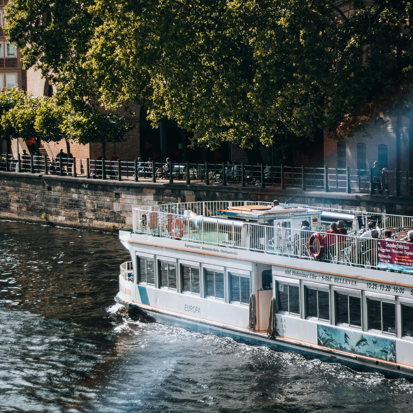 Berliner Sehenswürdigkeiten Personen auf einem Ausflugsboot auf einem Fluss neben einer Baumreihe und Gebäuden