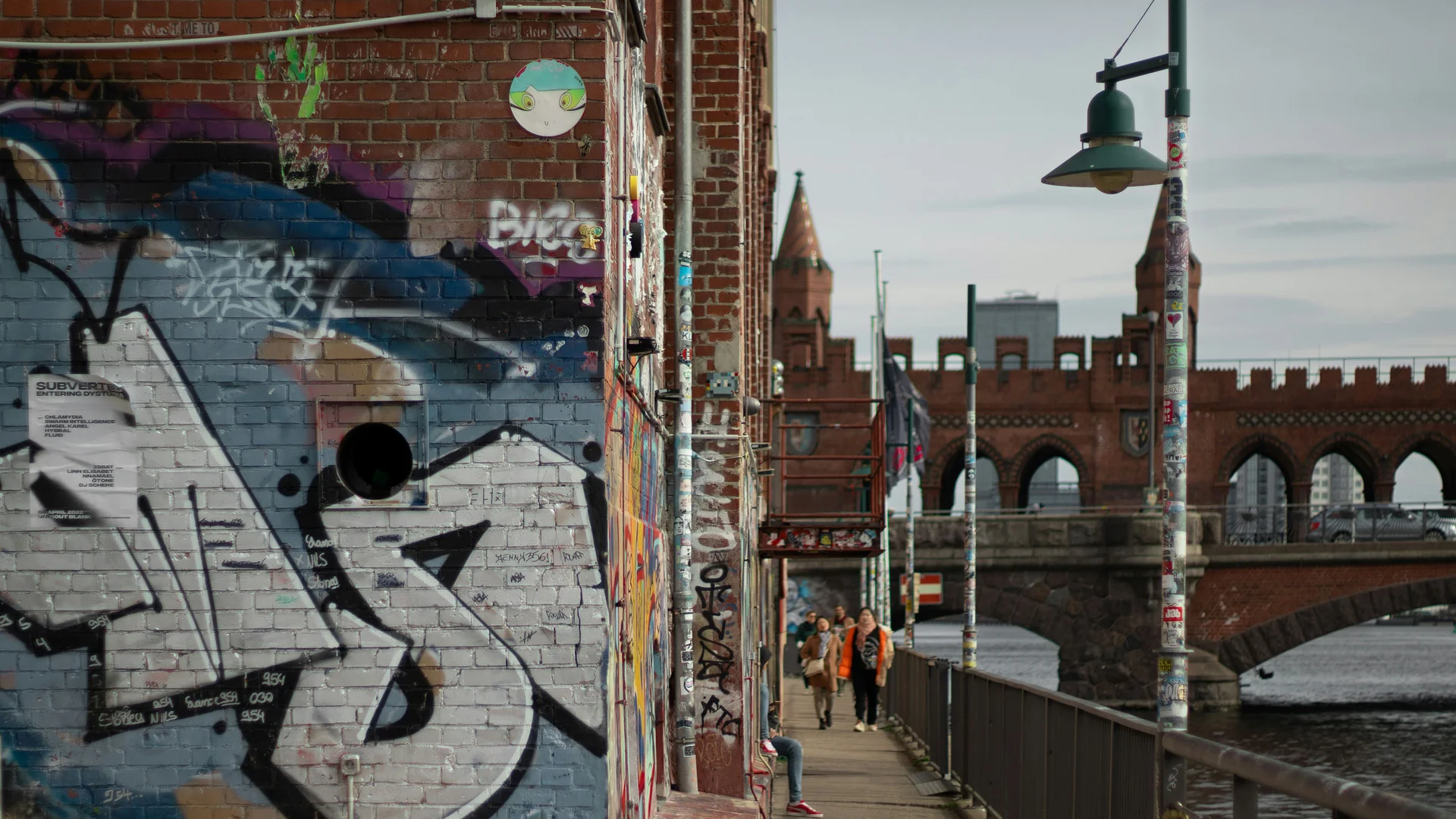 Bilder aus dem DASEYN Graffiti an Backsteinmauer neben Fluss mit Fußgängern und Brücke im Hintergrund