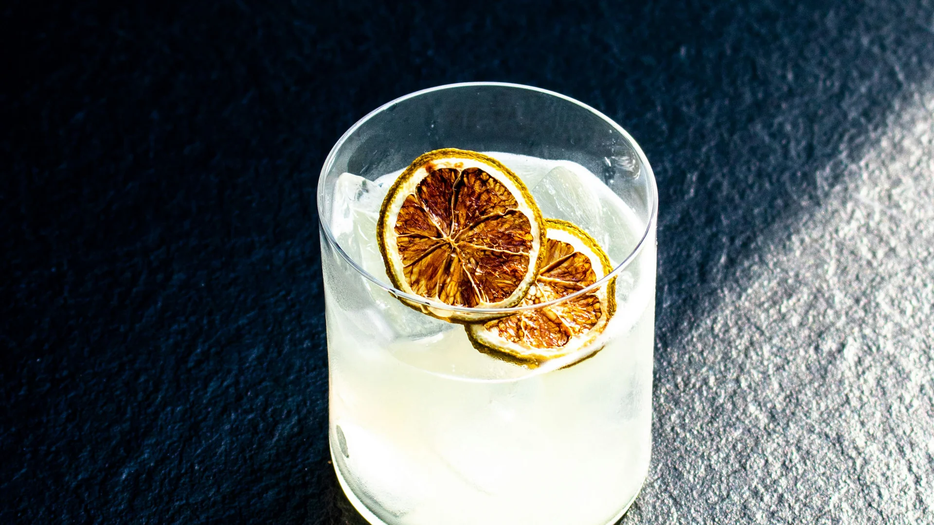 Bilder aus dem DASEYN Cocktail mit Eiswürfeln und getrockneten Limettenscheiben auf dunklem Untergrund